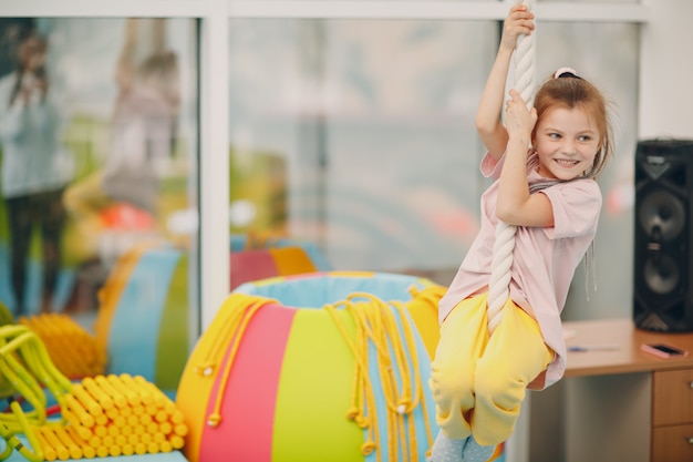 Kid girl doing exercises climbing tightrope in gym at kindergarten or elementary school. Children sport and fitness concept.