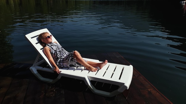 Kid girl lie on a sunbed in sunglasses and a boho silk shawl Child rest on a flood wood underwater pier The pavement is covered with water in the lake In the background are mountain and a forest