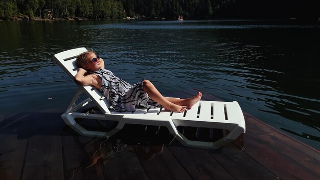 Kid girl lie on a sunbed in sunglasses and a boho silk shawl Child rest on a flood wood underwater pier The pavement is covered with water in the lake In the background are mountain and a forest