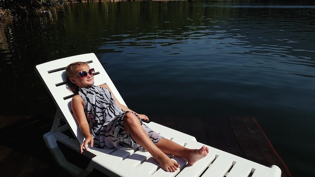 Kid girl lie on a sunbed in sunglasses and a boho silk shawl Child rest on a flood wood underwater pier The pavement is covered with water in lake