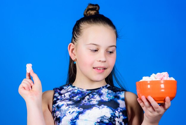 Kid girl with long hair likes sweets and treats Calorie and diet Hungry kid Girl smiling face hold bowl with sweet marshmallows in hand blue background Sweet and soft Sweet tooth concept