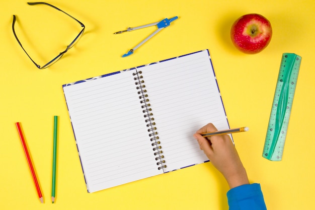 Kid hand with open notebook, pencils, glasses, fresh apple on yellow background