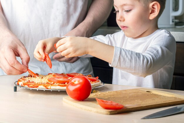 子供は父親が自家製ピザを調理するのを手伝います。検疫中の家族の娯楽。子供がピザを調理するプロセス。家にいて、社会的な距離を保ち、自己を孤立させる