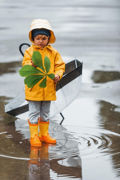 Kid in gele waterdichte mantellaarzen en met paraplu die buiten speelt na de regen