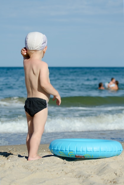 그의 뒤를 가진 해변에 수영 트렁크와 모자 서있는 아이가 거리를 찾습니다