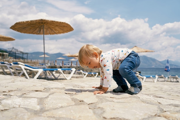 Kid is on all fours on a cobbled beach with sun loungers and\
umbrellas