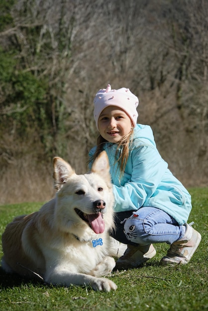 Kid is smiling sitting in a clearing and stroking a white domestic dog of mixed breed