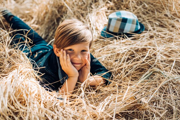 Kid jongen ligt op het hooi. Schattige kleine kind jongen met bladgoud op boerderij dorp achtergrond. Jongen in de hoed bereidt zich voor op zonnige herfstdag. Jeugd op de boerderij. Goede tijd in het dorp.