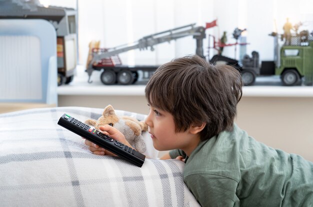 kid lying on a cushion holding tv remote control with toys in the background