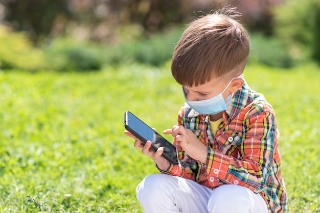 医療用マスクをかぶった子供が芝生に座って電話を見る