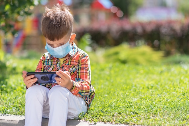 A kid in a medical mask sits on the grass and looks in the phone