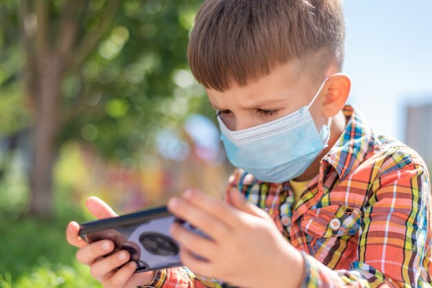 A kid in a medical mask sits on the grass and looks in the phone