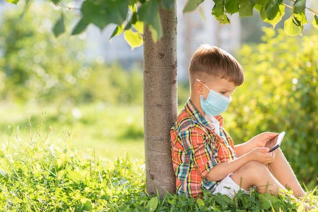 A kid in a medical mask sits on the grass and looks in the phone