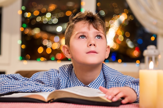 Kid met boek in de buurt van kaars. Kind wat betreft boek naast venster. Een wens doen met Kerstmis. Vakantie inspireert hem.
