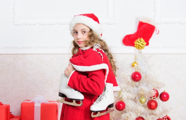 クリスマスツリーの近くの子供は冬のスケートを保持します小さな女の子はクリスマスプレゼントに満足しています最高の贈り物新年あけましておめでとうございます冬の休日フィギュアスケートのコンセプト冬の活動冬のレジャーのコンセプト