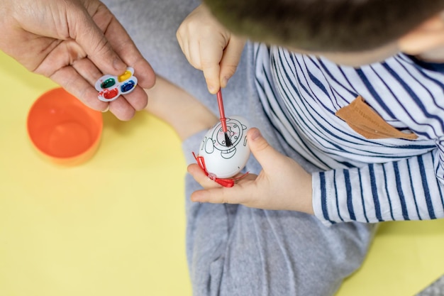 kid paints a easter egg with watercolor helped by the grandma easter holiday home activities