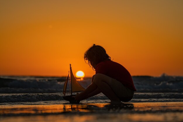 子供の少年の夏の日没時間の日没のシルエットで海のビーチでおもちゃのヨットで遊ぶ子供