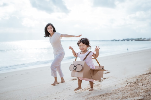 ビーチで走っている子供はお母さんと段ボールのおもちゃの飛行機で遊ぶ