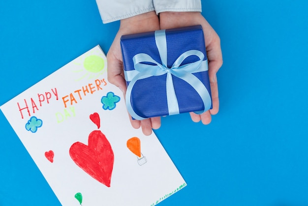 Kid's handen met geschenkdoos met tekening van het kind op blauw Concept Vaderdag wenskaart