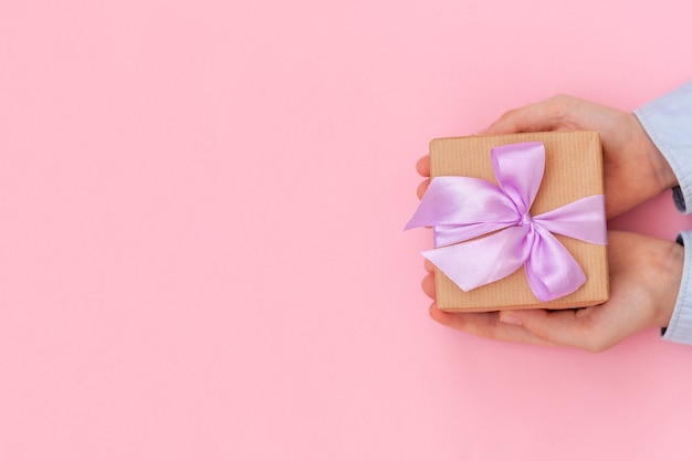 Kid's hands holding gift box wrapped in craft paper and tied with bow on pink wall.