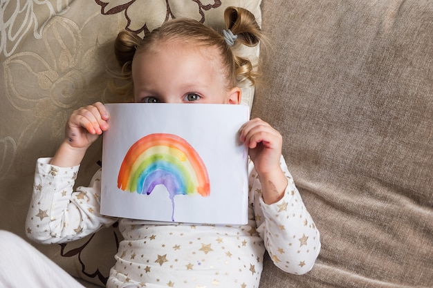 Kid schilderij regenboog tijdens covid-19. meisje dat een tekening met regenboog houdt. blijf thuis sociale mediacampagne voor coronavirus, laten we goed zijn, hoop tijdens pandemie