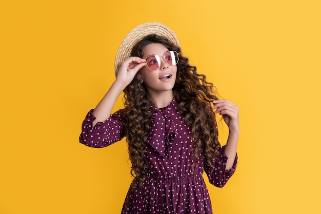 Kid shocked in straw hat and sunglasses with long brunette curly hair on yellow background