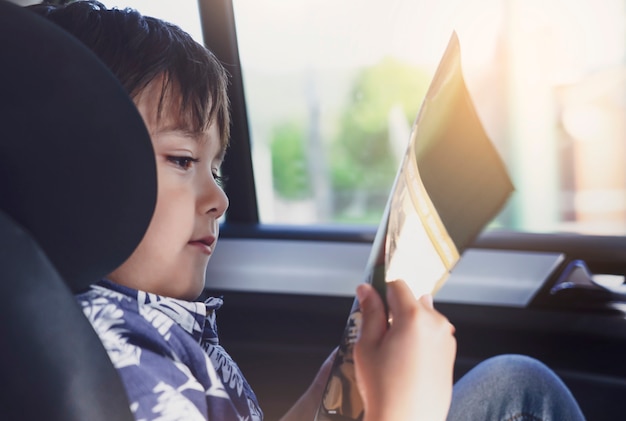 Kid siting on car seat and reading a book, Little boy sitting in the car in child safety seat, Portrait of toddler entertaining himserf on a road trip. Concept of safety taveling by car with children