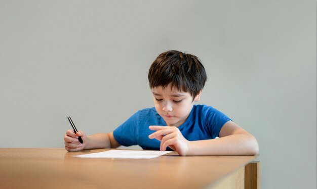 Kid siting on table doing homeworkChild boy holding black pen writing on white paperYoung boy practicing English words at home Elementary school and home schooling Distance Education concept