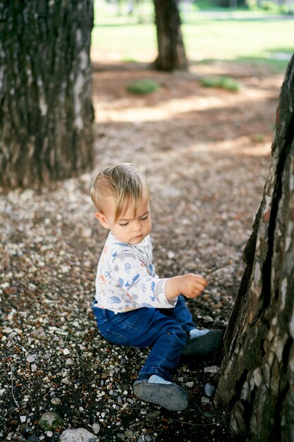 Kid sits on the ground near a large tree and holds a stick in his hand