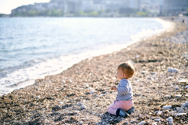 Kid sits on his knees on a pebble beach and looks at the sea