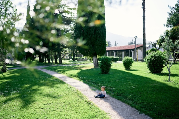 Kid sits on a path in the park among trees and greenery