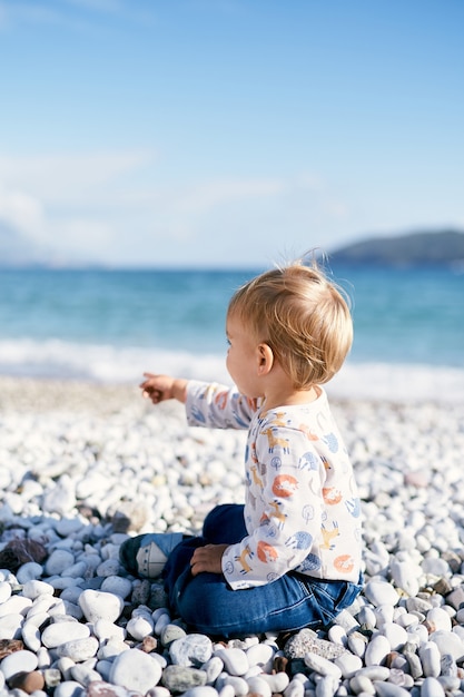 Il bambino si siede su una spiaggia di ciottoli e punta il dito verso il mare