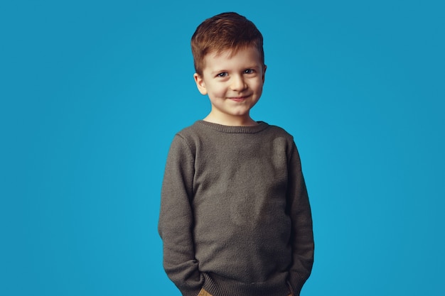 Kid smiling while holding hands in pockets isolated over blue background