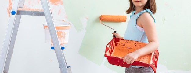 Kid standing with paint roller in hand, close-up. She is painting the wall. Redecoration, repair and repaint concept.