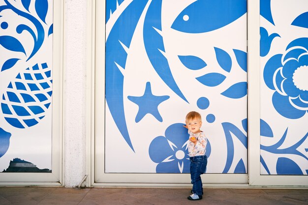 Kid stands against the background of blue flowers painted on a white wall