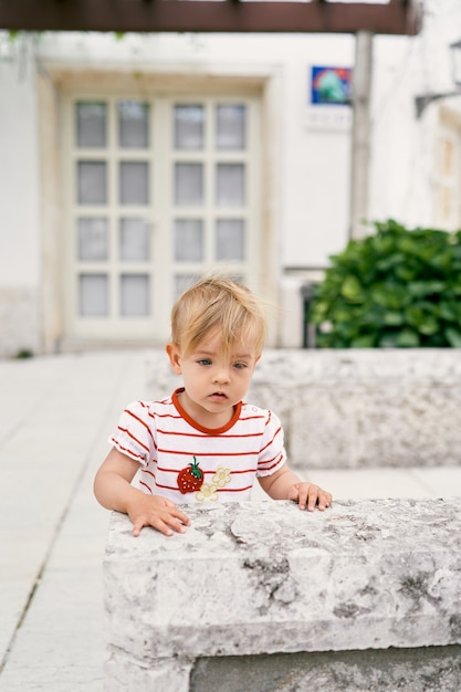 Kid stands at a stone fence in the courtyard of a house against the background of a glass door