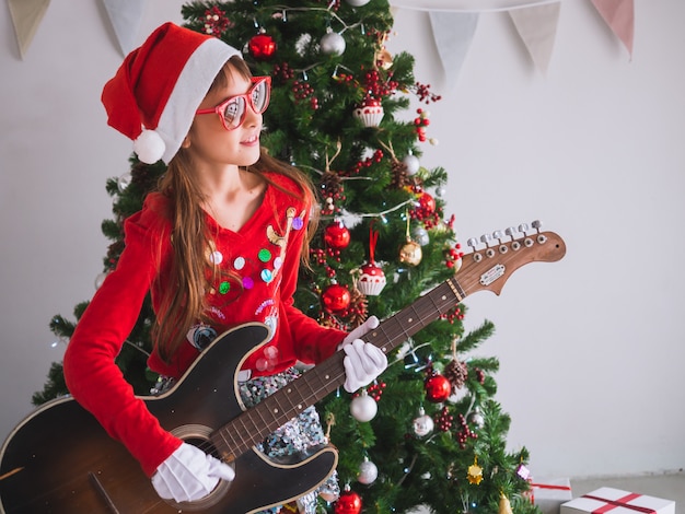 Foto kid viert kerstmis door de gitaar binnenshuis te tokkelen. een meisje speelt een lied met een glimlach op eerste kerstdag
