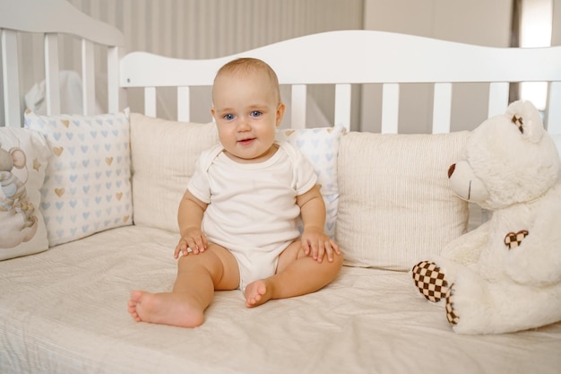 A kid in a white bodysuit in a white crib is sitting on his ass next to a teddy bear toy