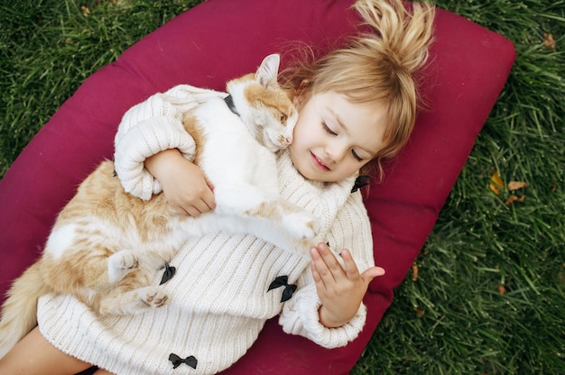Photo kid with cat lying on a blanket in the garden, caring for animals. child with kitty poses on backyard. happy childhood