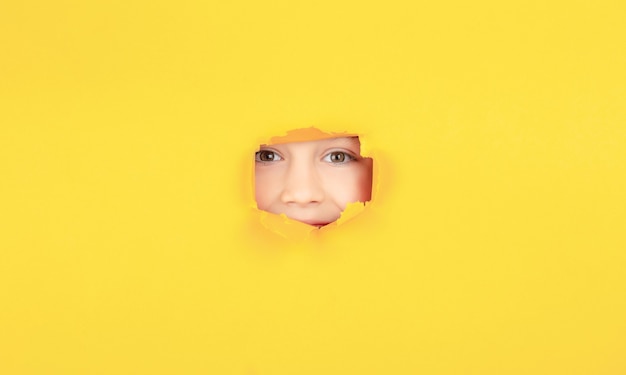 Kid with toothy smile shows face in paper hole. Positive child with toothy pleasant smile on face, keeps through torn hole in yellow paper. Breaking paper background. Emotions concept.