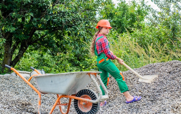 Kid working on construction site teen girl takes out rubble from wheelbarrow kid with shovel loading crushed stones Laying the foundation building project girl with wheelbarrow of rubble