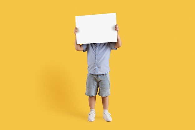 Photo kids ad full length shot of unrecognizable little boy covering his face with blank sheet of paper empty space