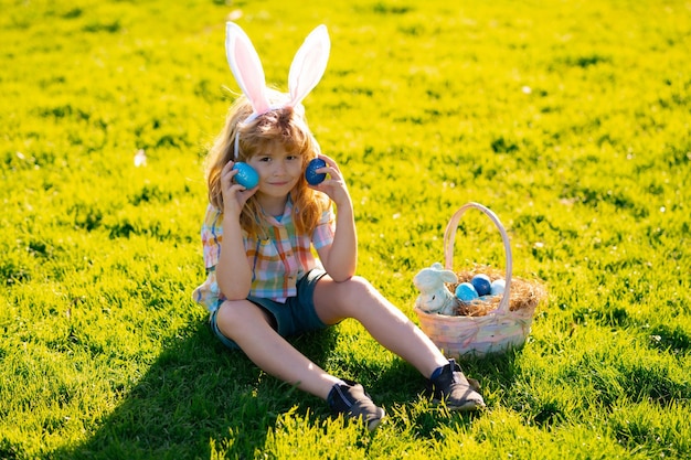 Мальчик в кроличьих ушах охотится за пасхальными яйцами на открытом воздухе Ребенок с пасхой в парке Охота за пасхальными яйцами