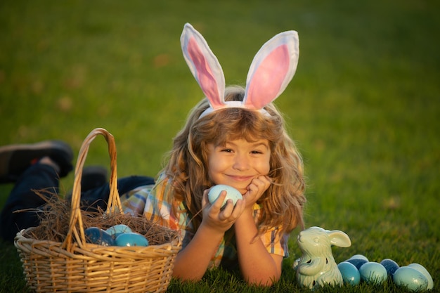 Детский мальчик охотится за пасхальными яйцами в парке, лежащим на траве. Кролики с кроличьими ушами. Крупным планом портрет милых детей.