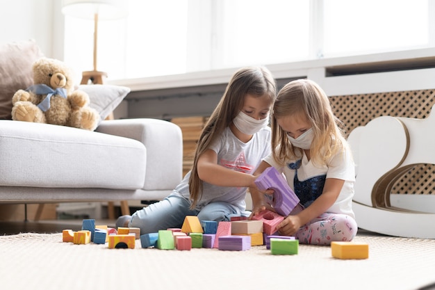 Bambini bambini che indossano una maschera per proteggere covid-19, giocando a giocattoli a blocchi nella stanza dei giochi. resta a casa in quarantena per prevenire la pandemia di coronavirus