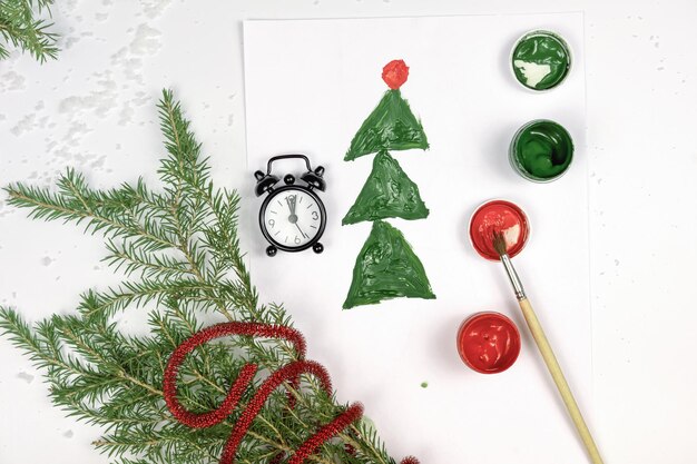 Foto bambini che disegnano un albero di natale e una sveglia con rami di albero di natale e colori su un tavolo bianco biglietto di auguri fatto a mano celebrazione del nuovo anno con attività per bambini vista dall'alto