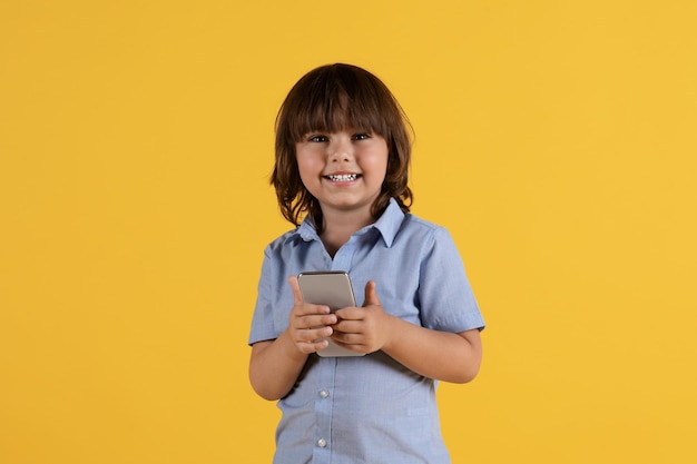 어린이 및 가제트 스마트폰을 들고 온라인 여유 공간에서 비디오를 보고 카메라에 미소 짓는 사랑스러운 작은 소년