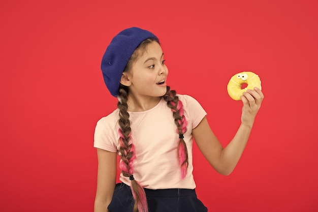 Kids huge fans of baked donuts Impossible to resist fresh made donut Girl hold glazed cute donut in hand red background Kid smiling girl ready to eat donut Sweets shop and bakery concept