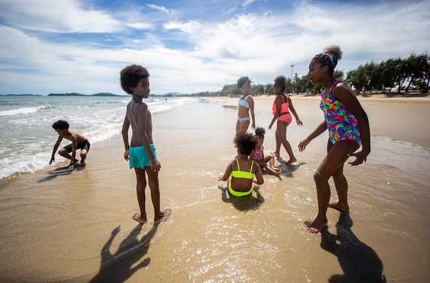 Bambini che giocano a correre sulla sabbia in spiaggia, un gruppo di bambini che si tengono per mano in fila sulla spiaggia in estate, vista posteriore contro il mare e il cielo blu