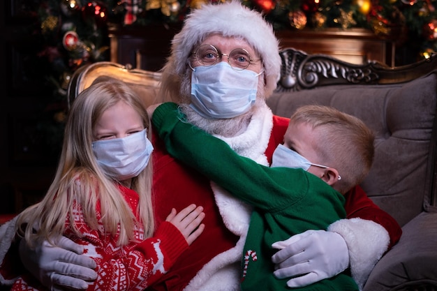 Kids and Santa in medical masks Portrait Girl boy and Santa Claus sitting near Christmas tree Happy New Year Covid 2021